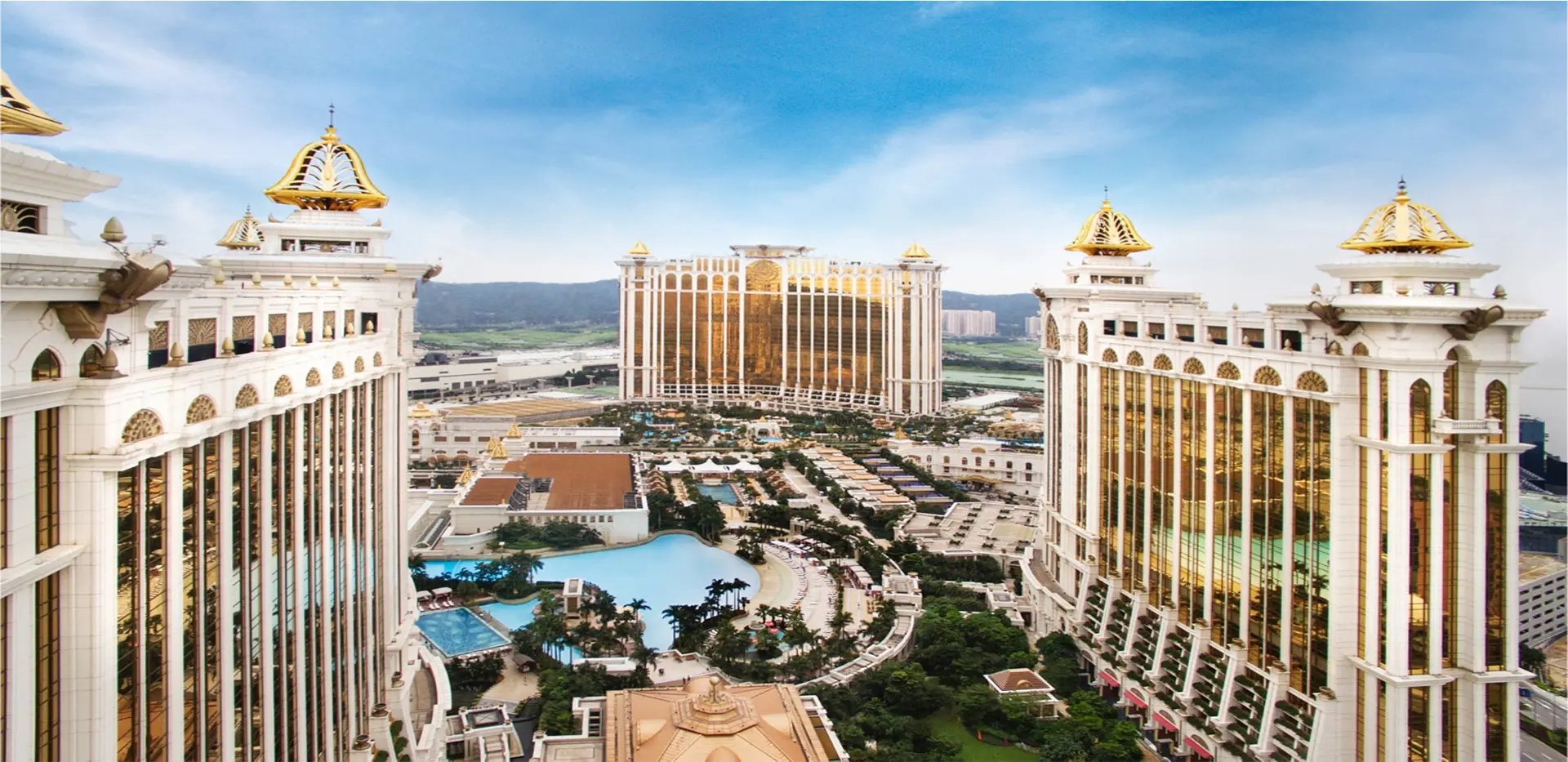 Kuvassa on henkeäsalpaava panoraamanäkymä valtavasta ja äärimmäisen ylellisestä lomakeskuksesta, joka tunnetaan nimellä Galaxy Macau. Kuvaa reunustavat etualalla kaksi massiivista, palatsimaista hotellitornia. Niiden vaaleaa julkisivua korostavat runsaat kultaiset yksityiskohdat, ja tornien huipulla kiiltävät näyttävät kultaiset kupolit. Tornien väliin jää laaja, trooppinen virkistysalue, jossa erottuu suuria uima-altaita, vehreitä puutarhoja ja palmuryhmiä. Taustalla kohoaa kolmas, kullanvärinen ja jättiläismäinen hotellirakennus. Näkymä huokuu "Aasian Las Vegasin" yltäkylläisyyttä ja luksusta.