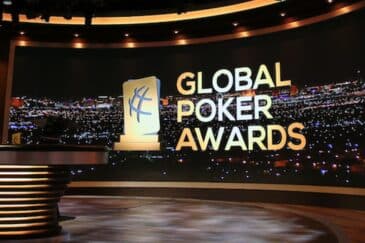 Kuvassa on näyttävä studioympäristö, jossa esitellään Global Poker Awards -gaalaa. Taustalla näkyy suuri, kaareva näyttö, joka esittää öistä kaupunkimaisemaa kirkkaine valoineen – todennäköisesti Las Vegasia. Näytön oikealla puolella on tapahtuman nimi “GLOBAL POKER AWARDS” suurin, vaaleankeltaisin kirjaimin. Nimen vieressä seisoo tyylitelty pysti-ikoni, joka kuvaa palkintoa. Etualalla on moderni, valaistu studiopöytä, jossa on pyöreitä, metallisia yksityiskohtia. Koko kuva huokuu juhlavuutta ja ammattimaisuutta, ja se korostaa pokerimaailman arvostetuimman palkintogaalan juhlallista tunnelmaa.