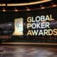 Kuvassa on näyttävä studioympäristö, jossa esitellään Global Poker Awards -gaalaa. Taustalla näkyy suuri, kaareva näyttö, joka esittää öistä kaupunkimaisemaa kirkkaine valoineen – todennäköisesti Las Vegasia. Näytön oikealla puolella on tapahtuman nimi “GLOBAL POKER AWARDS” suurin, vaaleankeltaisin kirjaimin. Nimen vieressä seisoo tyylitelty pysti-ikoni, joka kuvaa palkintoa. Etualalla on moderni, valaistu studiopöytä, jossa on pyöreitä, metallisia yksityiskohtia. Koko kuva huokuu juhlavuutta ja ammattimaisuutta, ja se korostaa pokerimaailman arvostetuimman palkintogaalan juhlallista tunnelmaa.