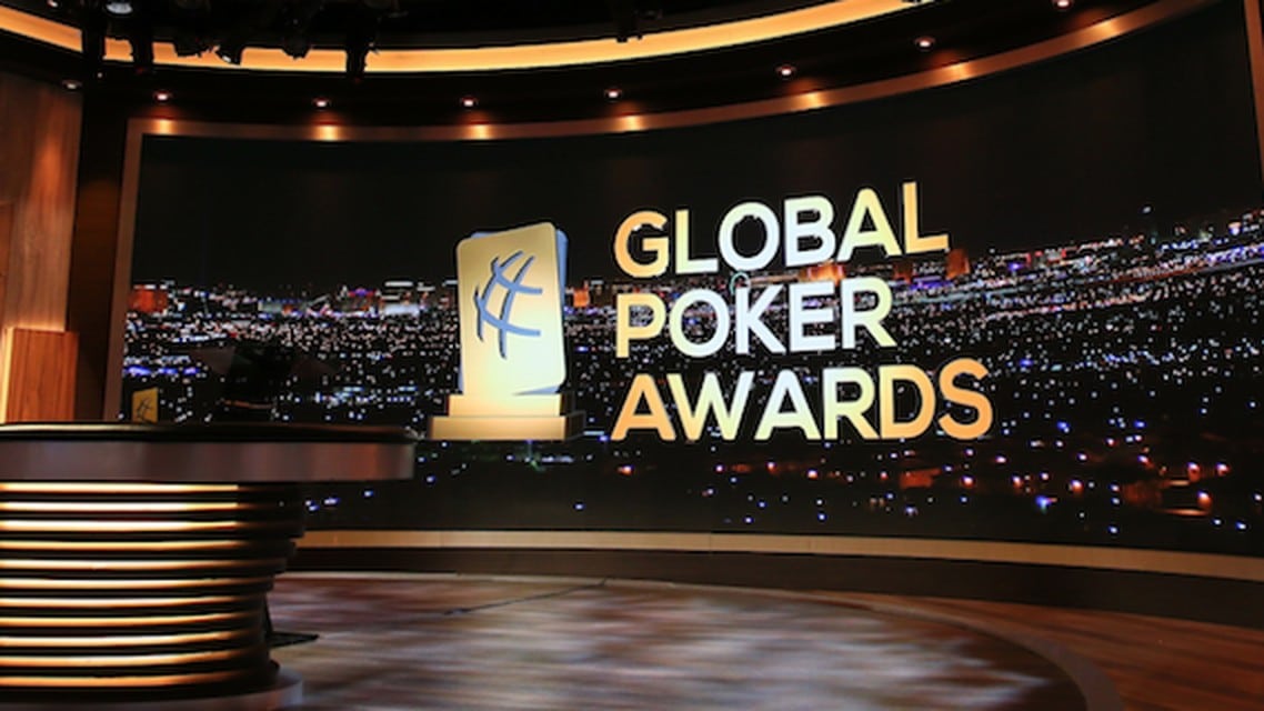 Kuvassa on näyttävä studioympäristö, jossa esitellään Global Poker Awards -gaalaa. Taustalla näkyy suuri, kaareva näyttö, joka esittää öistä kaupunkimaisemaa kirkkaine valoineen – todennäköisesti Las Vegasia. Näytön oikealla puolella on tapahtuman nimi “GLOBAL POKER AWARDS” suurin, vaaleankeltaisin kirjaimin. Nimen vieressä seisoo tyylitelty pysti-ikoni, joka kuvaa palkintoa. Etualalla on moderni, valaistu studiopöytä, jossa on pyöreitä, metallisia yksityiskohtia. Koko kuva huokuu juhlavuutta ja ammattimaisuutta, ja se korostaa pokerimaailman arvostetuimman palkintogaalan juhlallista tunnelmaa.