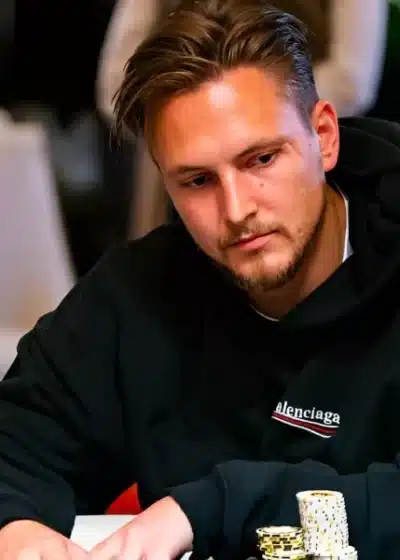 Kuvassa istuu nuori mies pokeripöydän ääressä keskittyneen ja hieman mietteliään näköisenä. Hän nojaa eteenpäin ja pitää käsiään pöydällä, jossa on useita siisteihin pinoihin järjestettyjä pelimerkkejä, pääosin keltaisia ja valkoisia. Miehellä on päällään musta huppari, jossa näkyy etupuolella pieni logomerkintä ja hihassa CoinPoker-tunnus. Tausta on pehmeästi sumea, mikä korostaa etualan tapahtumia ja luo ammattimaisen turnausympäristön. Valaistus on lämmin ja kohdistuu selkeästi pelaajaan, tuoden esiin hänen keskittyneen ilmeensä ja jännittyneen, pelitilanteeseen uppoutuneen tunnelman.