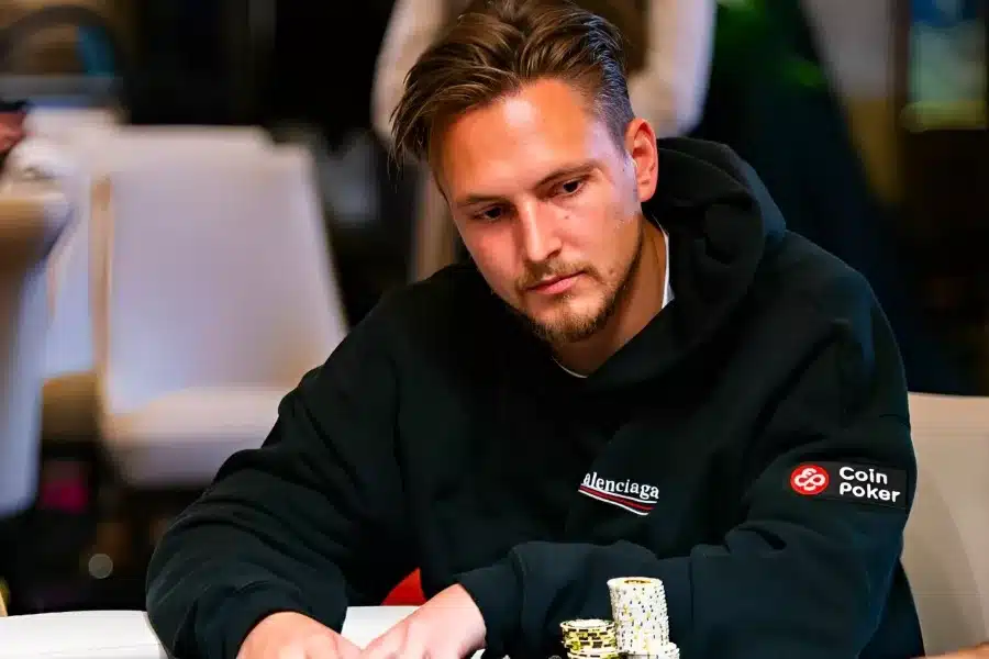Kuvassa istuu nuori mies pokeripöydän ääressä keskittyneen ja hieman mietteliään näköisenä. Hän nojaa eteenpäin ja pitää käsiään pöydällä, jossa on useita siisteihin pinoihin järjestettyjä pelimerkkejä, pääosin keltaisia ja valkoisia. Miehellä on päällään musta huppari, jossa näkyy etupuolella pieni logomerkintä ja hihassa CoinPoker-tunnus. Tausta on pehmeästi sumea, mikä korostaa etualan tapahtumia ja luo ammattimaisen turnausympäristön. Valaistus on lämmin ja kohdistuu selkeästi pelaajaan, tuoden esiin hänen keskittyneen ilmeensä ja jännittyneen, pelitilanteeseen uppoutuneen tunnelman.