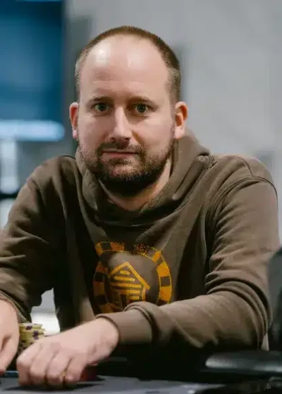 Kuvassa on mies istumassa pokeripöydän ääressä keskittyneenä. Hänellä on päällään ruskea huppari, jossa on keltainen graafinen kuvio keskellä. Edessään hänellä on pelimerkkejä ja pelikortteja, ja hänen kätensä lepäävät pöydällä valmiina seuraavaan siirtoon. Taustalla näkyy hämärästi valaistu pelisali, jossa on näyttöjä ja muita pelaajia. Kuva on otettu ammatillisesti ja se välittää rauhallisen mutta jännittyneen tunnelman, joka on tyypillinen pokeriturnauksen hetkille