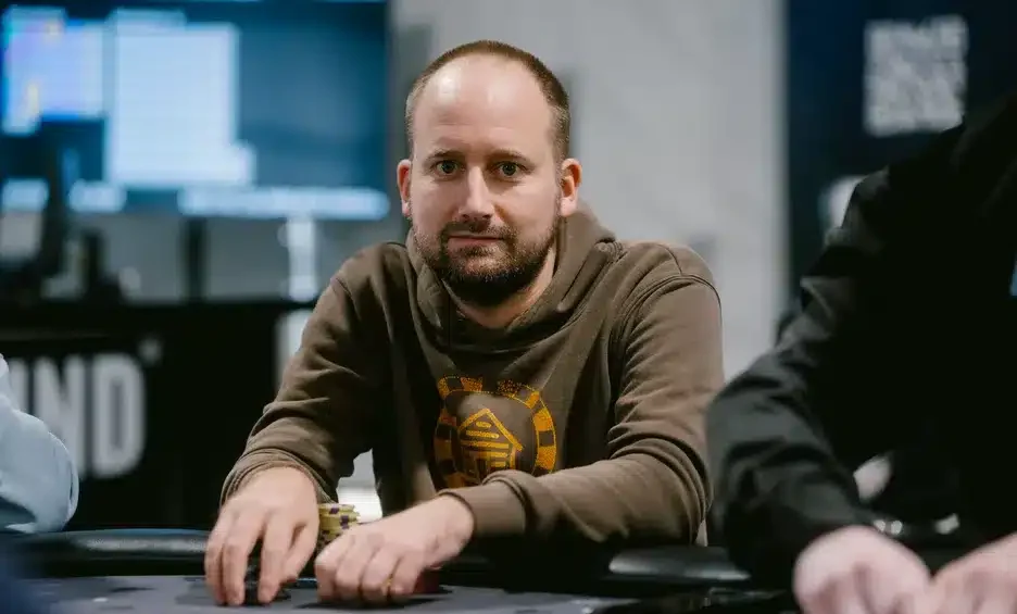 Kuvassa on mies istumassa pokeripöydän ääressä keskittyneenä. Hänellä on päällään ruskea huppari, jossa on keltainen graafinen kuvio keskellä. Edessään hänellä on pelimerkkejä ja pelikortteja, ja hänen kätensä lepäävät pöydällä valmiina seuraavaan siirtoon. Taustalla näkyy hämärästi valaistu pelisali, jossa on näyttöjä ja muita pelaajia. Kuva on otettu ammatillisesti ja se välittää rauhallisen mutta jännittyneen tunnelman, joka on tyypillinen pokeriturnauksen hetkille