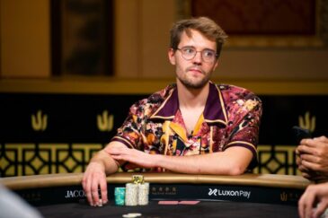 Kuvassa on nuori, silmälasipäinen mies, joka istuu pokeripöydän ääressä. Hänellä on yllään huomiota herättävä, värikäs ja kuviollinen kauluspaita. Miehen ilme on analyyttinen ja mietteliäs, ja hän katsoo sivuun samalla kun hänen kätensä lepää pöydällä pienten pelimerkkipinojen vieressä. Hän istuu tumman pokeripöydän ääressä, jossa näkyy Luxon Pay -sponsorilogo. Tausta on tyylikäs ja tumma, ja siinä erottuu kultaisia Triton Poker -logon koristeita, mikä viittaa korkean panoksen ammattilaisturnaukseen. Tunnelma on keskittynyt ja ammattimainen.