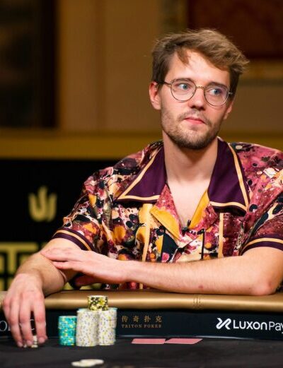 Kuvassa on nuori, silmälasipäinen mies, joka istuu pokeripöydän ääressä. Hänellä on yllään huomiota herättävä, värikäs ja kuviollinen kauluspaita. Miehen ilme on analyyttinen ja mietteliäs, ja hän katsoo sivuun samalla kun hänen kätensä lepää pöydällä pienten pelimerkkipinojen vieressä. Hän istuu tumman pokeripöydän ääressä, jossa näkyy Luxon Pay -sponsorilogo. Tausta on tyylikäs ja tumma, ja siinä erottuu kultaisia Triton Poker -logon koristeita, mikä viittaa korkean panoksen ammattilaisturnaukseen. Tunnelma on keskittynyt ja ammattimainen.