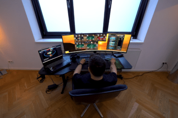 Kuvassa on nettipokerin pelaaja syventyneenä "grindaamiseen" modernin työpisteensä ääressä. Kuva on otettu yläviistosta pelaajan takaa, mikä antaa hyvän yleiskuvan hänen laitteistostaan. Pelaajan edessä on leveä, kaareva päänäyttö, jolle on järjestelty neljä samanaikaista pokeripöytää sekä oikeaan reunaan turnausaula tai tilastoikkuna. Vasemmalla puolella on lisäksi kannettava tietokone, jonka ruudulla on auki vielä yksi aktiivinen pelipöytä. Huone on sisustettu minimalistisesti, ja huomio kiinnittyy tyylikkääseen kalanruotoparkettiin sekä suureen kolmiosaiseen ikkunaan, joka tuo tilaan luonnonvaloa. Pelaajan keskittynyt asento ja monen pöydän samanaikainen hallinta viittaavat tavoitteelliseen tai ammattimaiseen pelaamiseen.