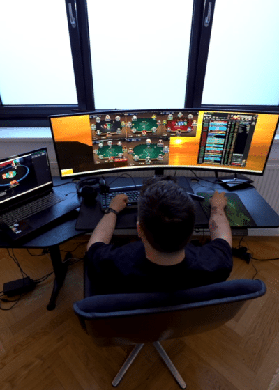 Kuvassa on nettipokerin pelaaja syventyneenä "grindaamiseen" modernin työpisteensä ääressä. Kuva on otettu yläviistosta pelaajan takaa, mikä antaa hyvän yleiskuvan hänen laitteistostaan. Pelaajan edessä on leveä, kaareva päänäyttö, jolle on järjestelty neljä samanaikaista pokeripöytää sekä oikeaan reunaan turnausaula tai tilastoikkuna. Vasemmalla puolella on lisäksi kannettava tietokone, jonka ruudulla on auki vielä yksi aktiivinen pelipöytä. Huone on sisustettu minimalistisesti, ja huomio kiinnittyy tyylikkääseen kalanruotoparkettiin sekä suureen kolmiosaiseen ikkunaan, joka tuo tilaan luonnonvaloa. Pelaajan keskittynyt asento ja monen pöydän samanaikainen hallinta viittaavat tavoitteelliseen tai ammattimaiseen pelaamiseen.