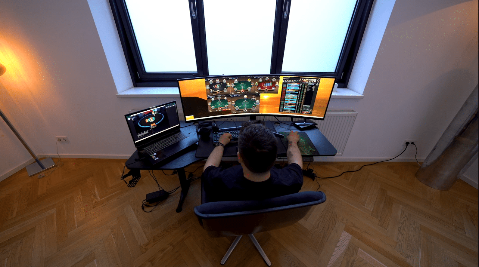 Kuvassa on nettipokerin pelaaja syventyneenä "grindaamiseen" modernin työpisteensä ääressä. Kuva on otettu yläviistosta pelaajan takaa, mikä antaa hyvän yleiskuvan hänen laitteistostaan. Pelaajan edessä on leveä, kaareva päänäyttö, jolle on järjestelty neljä samanaikaista pokeripöytää sekä oikeaan reunaan turnausaula tai tilastoikkuna. Vasemmalla puolella on lisäksi kannettava tietokone, jonka ruudulla on auki vielä yksi aktiivinen pelipöytä. Huone on sisustettu minimalistisesti, ja huomio kiinnittyy tyylikkääseen kalanruotoparkettiin sekä suureen kolmiosaiseen ikkunaan, joka tuo tilaan luonnonvaloa. Pelaajan keskittynyt asento ja monen pöydän samanaikainen hallinta viittaavat tavoitteelliseen tai ammattimaiseen pelaamiseen.