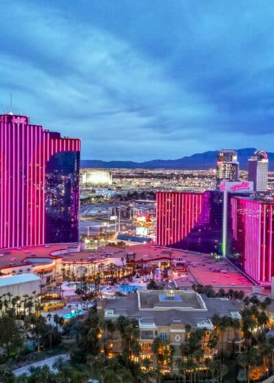 Kuvassa on laaja iltahämärän panoraama Las Vegasin kaupunkimaisemasta, jonka keskiössä on Palms Casino Resort -hotellikompleksi. Kuvaa hallitsevat Palmsin kolme suurta hotellitornia, jotka on valaistu näyttävällä ja kirkkaalla pinkillä neonvalolla. Erityisesti vasemmalla oleva päärakennus ja oikealla olevat kaksi pienempää tornia hehkuvat voimakkaasti sinistä taivasta vasten. Etualalla näkyy hotellin laajempi, matalampi osa sekä ylellinen uima-allasalue, jota reunustavat palmut ja huvilamaiset rakennukset. Taustalla levittäytyy koko Las Vegasin laakso täynnä valoja, ja kaukaisuudessa siintävät vuorten tummat siluetit. Kuva on otettu juuri auringonlaskun jälkeen, ja se vangitsee täydellisesti Las Vegasin energisen ja loisteliaan tunnelman.