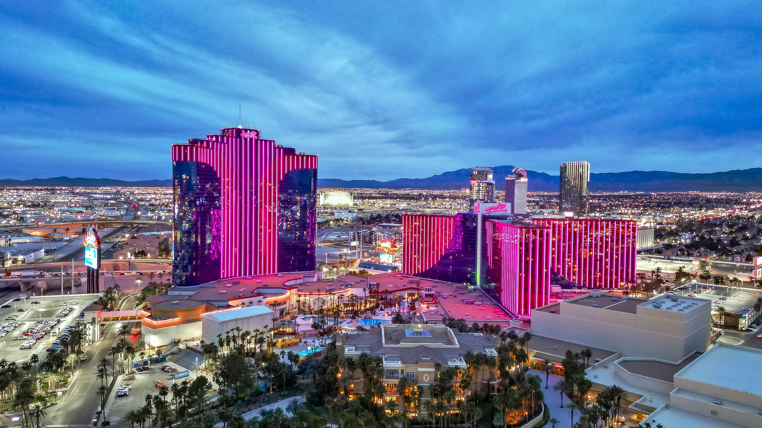 Kuvassa on laaja iltahämärän panoraama Las Vegasin kaupunkimaisemasta, jonka keskiössä on Palms Casino Resort -hotellikompleksi. Kuvaa hallitsevat Palmsin kolme suurta hotellitornia, jotka on valaistu näyttävällä ja kirkkaalla pinkillä neonvalolla. Erityisesti vasemmalla oleva päärakennus ja oikealla olevat kaksi pienempää tornia hehkuvat voimakkaasti sinistä taivasta vasten. Etualalla näkyy hotellin laajempi, matalampi osa sekä ylellinen uima-allasalue, jota reunustavat palmut ja huvilamaiset rakennukset. Taustalla levittäytyy koko Las Vegasin laakso täynnä valoja, ja kaukaisuudessa siintävät vuorten tummat siluetit. Kuva on otettu juuri auringonlaskun jälkeen, ja se vangitsee täydellisesti Las Vegasin energisen ja loisteliaan tunnelman.