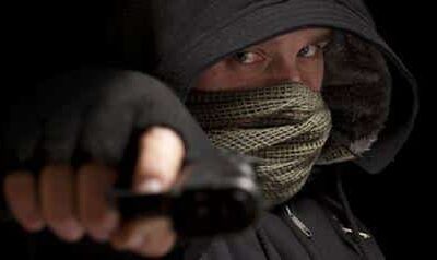 Kuvassa näkyy tummiin vaatteisiin pukeutunut henkilö, jonka olemus on uhkaava ja tarkoituksella peitetty. Hänellä on yllään musta huppu, joka varjostaa kasvoja, sekä kuviollinen huivi tai maski, joka peittää suun ja osan poskista. Vain silmät näkyvät selvästi, ja katse on intensiivinen ja suoraan kohti kameraa. Henkilö ojentaa kätensä eteenpäin ja pitelee siinä esinettä, joka muistuttaa asetta, luoden tilanteeseen jännittyneen ja vaarallisen tunnelman. Tausta on täysin musta, mikä korostaa hahmoa ja lisää kuvan dramaattisuutta sekä anonymiteetin tuntua.