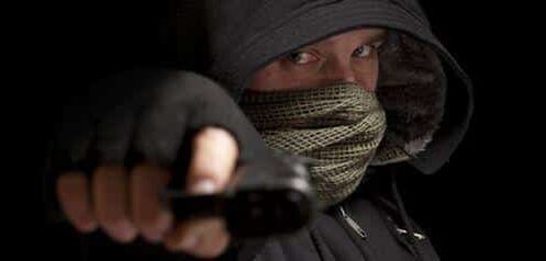 Kuvassa näkyy tummiin vaatteisiin pukeutunut henkilö, jonka olemus on uhkaava ja tarkoituksella peitetty. Hänellä on yllään musta huppu, joka varjostaa kasvoja, sekä kuviollinen huivi tai maski, joka peittää suun ja osan poskista. Vain silmät näkyvät selvästi, ja katse on intensiivinen ja suoraan kohti kameraa. Henkilö ojentaa kätensä eteenpäin ja pitelee siinä esinettä, joka muistuttaa asetta, luoden tilanteeseen jännittyneen ja vaarallisen tunnelman. Tausta on täysin musta, mikä korostaa hahmoa ja lisää kuvan dramaattisuutta sekä anonymiteetin tuntua.