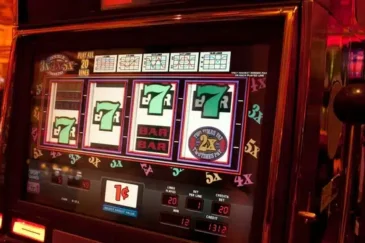 Kuvassa on peliautomaatti, jonka kirkkaasti valaistu näyttö esittää klassista kolikkopeliä. Ruudulla näkyy neljä rullaa, joista kolmessa on vihreä numero seitsemän ja yhdessä “BAR”-symboli sekä “2x”-kerroin. Pelin teema on perinteinen kasino-tyylinen, ja sen ympärillä loistaa lämmin, punertava valaistus, joka luo tunnelmallisen ja houkuttelevan ilmapiirin. Näytön alareunassa on tietoja panoksesta, voitoista ja krediiteistä, ja kolikoiden arvo on yksi sentti. Oikealla puolella näkyy metallinen pelivipu. Kuva välittää Las Vegas -tyylistä jännitystä ja pelaamisen odotuksen tunnetta.