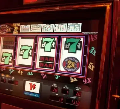 Kuvassa on peliautomaatti, jonka kirkkaasti valaistu näyttö esittää klassista kolikkopeliä. Ruudulla näkyy neljä rullaa, joista kolmessa on vihreä numero seitsemän ja yhdessä “BAR”-symboli sekä “2x”-kerroin. Pelin teema on perinteinen kasino-tyylinen, ja sen ympärillä loistaa lämmin, punertava valaistus, joka luo tunnelmallisen ja houkuttelevan ilmapiirin. Näytön alareunassa on tietoja panoksesta, voitoista ja krediiteistä, ja kolikoiden arvo on yksi sentti. Oikealla puolella näkyy metallinen pelivipu. Kuva välittää Las Vegas -tyylistä jännitystä ja pelaamisen odotuksen tunnetta.