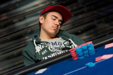 Kuvassa nuori mies on nukahtanut pokeripöydän ääreen. Hän istuu nojautuneena taaksepäin, pää kallellaan sivulle, silmät kiinni. Miehellä on päässään punainen lippalakki ja yllään tummanvihreä huppari, jonka alla näkyy harmaa t-paita. Hänen edessään pöydällä on siistissä pinossa sinisiä ja punaisia pelimerkkejä sekä muutama pelikortti. Tausta on epätarkka ja tummasävyinen, mikä korostaa etualan tapahtumaa. Kuva välittää väsymyksen ja pitkän pelisession tunnelman, joka on tuttu monille pokeriturnauksiin osallistuville pelaajille.