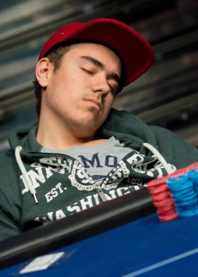 Kuvassa nuori mies on nukahtanut pokeripöydän ääreen. Hän istuu nojautuneena taaksepäin, pää kallellaan sivulle, silmät kiinni. Miehellä on päässään punainen lippalakki ja yllään tummanvihreä huppari, jonka alla näkyy harmaa t-paita. Hänen edessään pöydällä on siistissä pinossa sinisiä ja punaisia pelimerkkejä sekä muutama pelikortti. Tausta on epätarkka ja tummasävyinen, mikä korostaa etualan tapahtumaa. Kuva välittää väsymyksen ja pitkän pelisession tunnelman, joka on tuttu monille pokeriturnauksiin osallistuville pelaajille.