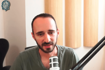 Kuvassa on mies, joka istuu mikrofonin edessä ja näyttää puhuvan, mahdollisesti podcastissa tai videokeskustelussa. Hänellä on lyhyet, tummat hiukset ja parta, ja hän pukeutuu vihreään V-kaulaiseen T-paitaan. Taustalla näkyy vaalea seinä ja pystysuorassa asennossa olevia puupaneeleja, jotka todennäköisesti toimivat ääntä vaimentavina elementteinä. Vasemmalla puolella on musta toimistotuoli ja oikealla suuri mikrofonivarsi, joka on kiinnitetty pöytään. Kuvan vasempaan yläkulmaan on lisätty logomerkki, jossa on ihmisen pään muotoinen kuvio rataskomponentteineen. Tunnelma on rauhallinen ja ammattimainen.