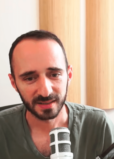 Kuvassa on mies, joka istuu mikrofonin edessä ja näyttää puhuvan, mahdollisesti podcastissa tai videokeskustelussa. Hänellä on lyhyet, tummat hiukset ja parta, ja hän pukeutuu vihreään V-kaulaiseen T-paitaan. Taustalla näkyy vaalea seinä ja pystysuorassa asennossa olevia puupaneeleja, jotka todennäköisesti toimivat ääntä vaimentavina elementteinä. Vasemmalla puolella on musta toimistotuoli ja oikealla suuri mikrofonivarsi, joka on kiinnitetty pöytään. Kuvan vasempaan yläkulmaan on lisätty logomerkki, jossa on ihmisen pään muotoinen kuvio rataskomponentteineen. Tunnelma on rauhallinen ja ammattimainen.