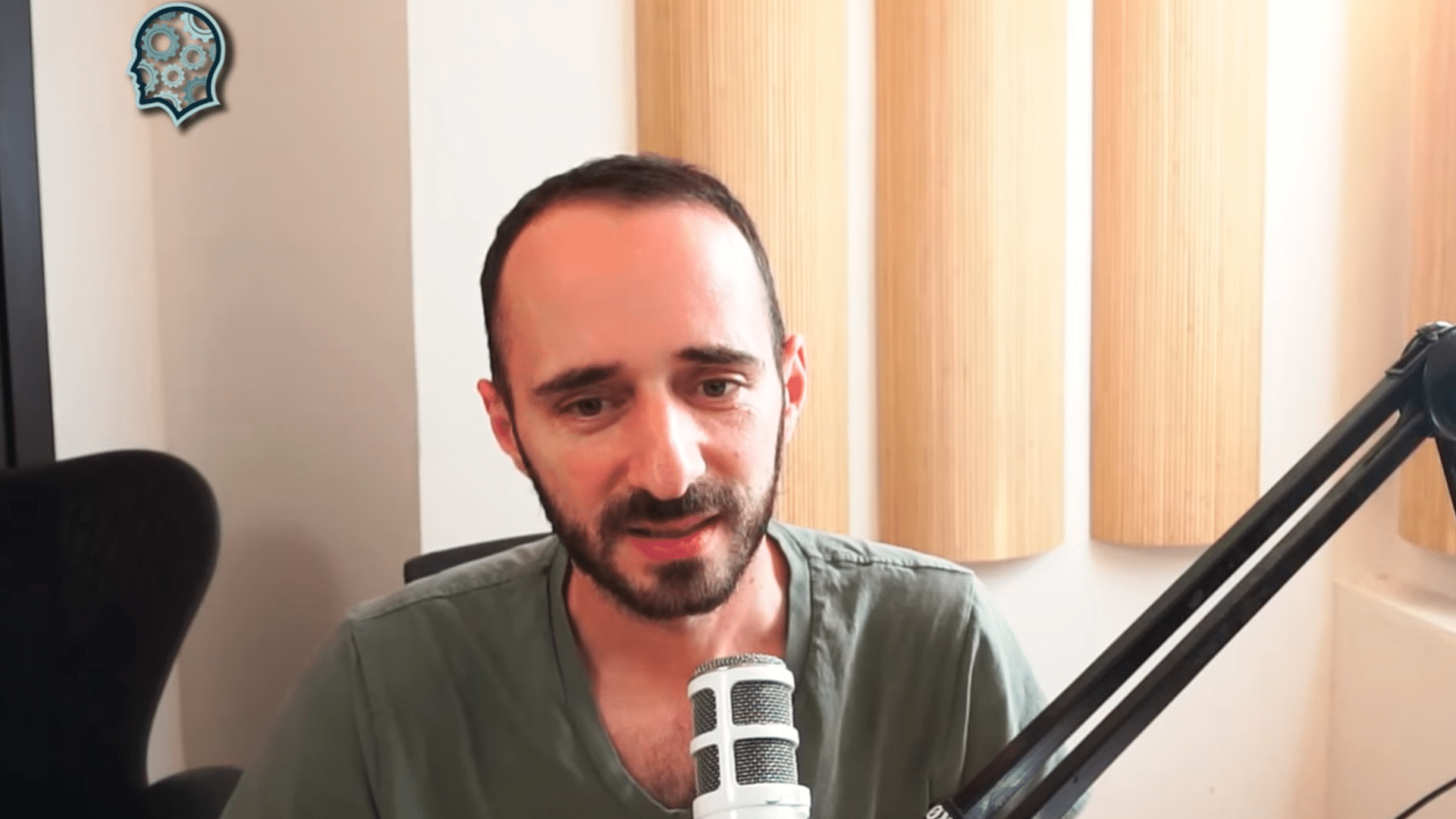 Kuvassa on mies, joka istuu mikrofonin edessä ja näyttää puhuvan, mahdollisesti podcastissa tai videokeskustelussa. Hänellä on lyhyet, tummat hiukset ja parta, ja hän pukeutuu vihreään V-kaulaiseen T-paitaan. Taustalla näkyy vaalea seinä ja pystysuorassa asennossa olevia puupaneeleja, jotka todennäköisesti toimivat ääntä vaimentavina elementteinä. Vasemmalla puolella on musta toimistotuoli ja oikealla suuri mikrofonivarsi, joka on kiinnitetty pöytään. Kuvan vasempaan yläkulmaan on lisätty logomerkki, jossa on ihmisen pään muotoinen kuvio rataskomponentteineen. Tunnelma on rauhallinen ja ammattimainen.