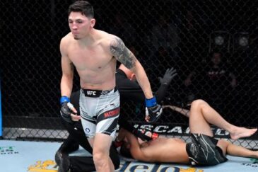 Kuvassa on dramaattinen hetki UFC-vapaaotteluillasta. Kuvan etualalla seisoo ottelun voittaja, jolla on yllään valkoiset ottelushortsit ja tatuointeja vasemmassa käsivarressaan. Hänen ilmeensä on intensiivinen ja latautunut, kun hän katsoo juuri voittamaansa vastustajaa. Hänen vastustajansa makaa selällään kanveesissa häkin reunalla, ilmeisesti tyrmättynä ottelun päätyttyä. Taustalla näkyy häkin verkkoaita ja 'gopuff'-sponsorilogo. Kuva vangitsee täydellisesti tyrmäysvoiton välittömän reaktion ja raa'an, urheilullisen hetken.