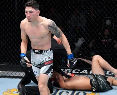 Kuvassa on dramaattinen hetki UFC-vapaaotteluillasta. Kuvan etualalla seisoo ottelun voittaja, jolla on yllään valkoiset ottelushortsit ja tatuointeja vasemmassa käsivarressaan. Hänen ilmeensä on intensiivinen ja latautunut, kun hän katsoo juuri voittamaansa vastustajaa. Hänen vastustajansa makaa selällään kanveesissa häkin reunalla, ilmeisesti tyrmättynä ottelun päätyttyä. Taustalla näkyy häkin verkkoaita ja 'gopuff'-sponsorilogo. Kuva vangitsee täydellisesti tyrmäysvoiton välittömän reaktion ja raa'an, urheilullisen hetken.