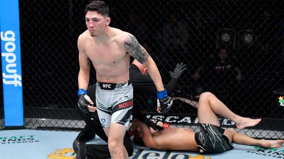 Kuvassa on dramaattinen hetki UFC-vapaaotteluillasta. Kuvan etualalla seisoo ottelun voittaja, jolla on yllään valkoiset ottelushortsit ja tatuointeja vasemmassa käsivarressaan. Hänen ilmeensä on intensiivinen ja latautunut, kun hän katsoo juuri voittamaansa vastustajaa. Hänen vastustajansa makaa selällään kanveesissa häkin reunalla, ilmeisesti tyrmättynä ottelun päätyttyä. Taustalla näkyy häkin verkkoaita ja 'gopuff'-sponsorilogo. Kuva vangitsee täydellisesti tyrmäysvoiton välittömän reaktion ja raa'an, urheilullisen hetken.