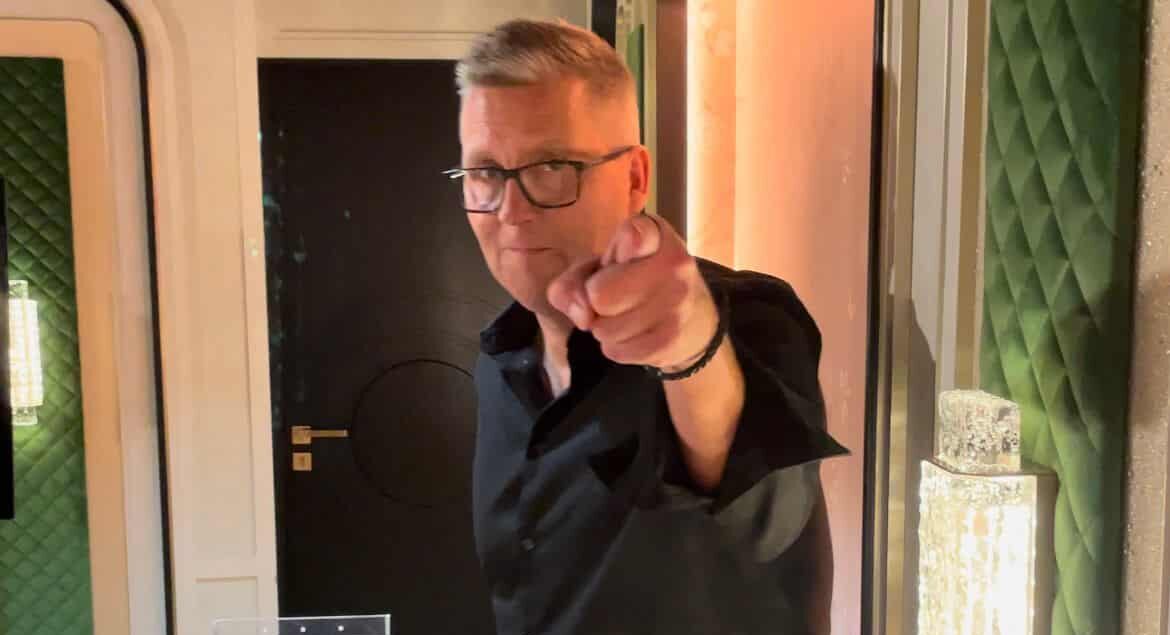 Jani Sievinen osoittaa sormella. Hänellä on päällään musta kauluspaita. Takana näkyy tumma ovi ja vaaleaa seinää.