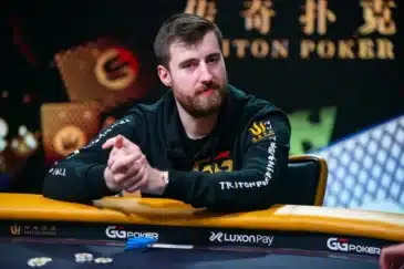 Kuvassa istuu pokeripöydän ääressä henkilö, joka vaikuttaa keskittyneeltä ja rauhalliselta. Hänellä on tummat, urheilulliset vaatteet ja huppari, jossa näkyy useita logomerkintöjä sekä Triton Poker -brändi. Taustalla on näyttävä, tummasävyinen lava tai studioympäristö, jossa näkyy koristeellisia valoja ja pokeriturnausta mainostavia tekstejä. Henkilö nojaa käsillään pöytään ja pitää käsiään lomittain, ikään kuin odottaisi seuraavaa siirtoa tai miettisi pelitilannetta. Pokeripöydällä näkyy pelimerkkejä ja varusteita, jotka korostavat kilpailullista ilmapiiriä. Koko kuva välittää ammattimaisen ja vakavan tunnelman, joka on tyypillinen korkean tason pokeritapahtumille.