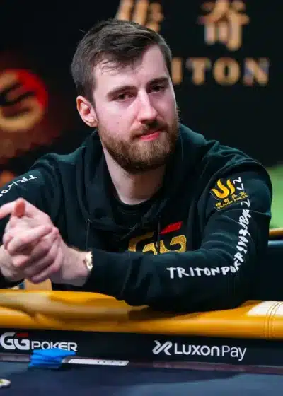 Kuvassa istuu pokeripöydän ääressä henkilö, joka vaikuttaa keskittyneeltä ja rauhalliselta. Hänellä on tummat, urheilulliset vaatteet ja huppari, jossa näkyy useita logomerkintöjä sekä Triton Poker -brändi. Taustalla on näyttävä, tummasävyinen lava tai studioympäristö, jossa näkyy koristeellisia valoja ja pokeriturnausta mainostavia tekstejä. Henkilö nojaa käsillään pöytään ja pitää käsiään lomittain, ikään kuin odottaisi seuraavaa siirtoa tai miettisi pelitilannetta. Pokeripöydällä näkyy pelimerkkejä ja varusteita, jotka korostavat kilpailullista ilmapiiriä. Koko kuva välittää ammattimaisen ja vakavan tunnelman, joka on tyypillinen korkean tason pokeritapahtumille.