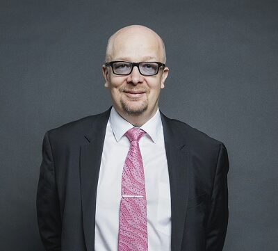 Aki Pyysing seisoo hymyillen tumma puku päällään.Puvun alla on valkoinen kauluspaita ja pinkki kravatti. Tausta on harmaata.