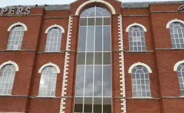 Kuvassa on suuri, kolmikerroksinen punatiilinen rakennus, jonka katonrajassa lukee valkoisilla kirjaimilla "ASPERS". Kyseessä on Aspers-kasino. Kuvan etualalla, suoraan rakennuksen edessä, on pysäköitynä brittiläinen, sini-keltainen poliisiauto. Huomio kiinnittyy rakennuksen moderniin lasiseen pääsisäänkäyntiin, joka on selvästi vaurioitunut. Oviaukko on rikki, ja sen edustalla maassa on puunsirpaleita ja laudanpätkiä, mikä viittaa siihen, että paikalla on tapahtunut jonkinlainen välikohtaus, kuten murto tai törmäys. Harmaa ja pilvinen taivas luo vakavan tunnelman.