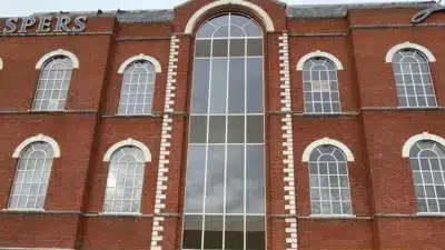 Kuvassa on suuri, kolmikerroksinen punatiilinen rakennus, jonka katonrajassa lukee valkoisilla kirjaimilla "ASPERS". Kyseessä on Aspers-kasino. Kuvan etualalla, suoraan rakennuksen edessä, on pysäköitynä brittiläinen, sini-keltainen poliisiauto. Huomio kiinnittyy rakennuksen moderniin lasiseen pääsisäänkäyntiin, joka on selvästi vaurioitunut. Oviaukko on rikki, ja sen edustalla maassa on puunsirpaleita ja laudanpätkiä, mikä viittaa siihen, että paikalla on tapahtunut jonkinlainen välikohtaus, kuten murto tai törmäys. Harmaa ja pilvinen taivas luo vakavan tunnelman.