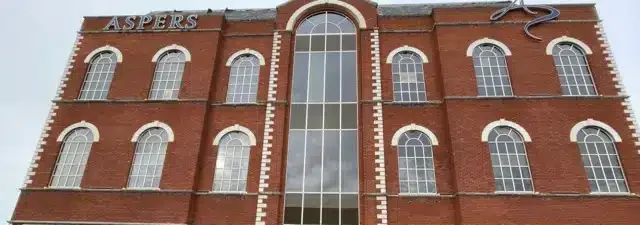 Kuvassa on suuri, kolmikerroksinen punatiilinen rakennus, jonka katonrajassa lukee valkoisilla kirjaimilla "ASPERS". Kyseessä on Aspers-kasino. Kuvan etualalla, suoraan rakennuksen edessä, on pysäköitynä brittiläinen, sini-keltainen poliisiauto. Huomio kiinnittyy rakennuksen moderniin lasiseen pääsisäänkäyntiin, joka on selvästi vaurioitunut. Oviaukko on rikki, ja sen edustalla maassa on puunsirpaleita ja laudanpätkiä, mikä viittaa siihen, että paikalla on tapahtunut jonkinlainen välikohtaus, kuten murto tai törmäys. Harmaa ja pilvinen taivas luo vakavan tunnelman.