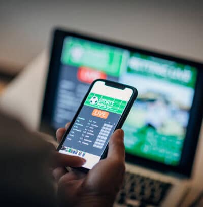 Vedonlyöntisivusto auki älypuhelimella ja taustalla pöyrii urheilulähetys läppärin ruudussa