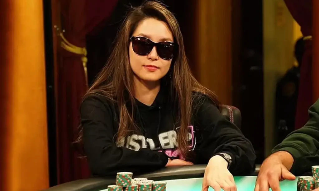 Nainen istuu pokeripöydässä. Hänellä on päällään musta huppari ja mustat aurinkolasit. Pokeripöydällä on paljon pelimerkkejä.