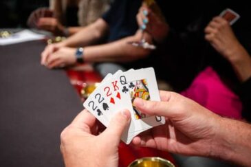 Kädessä on viisi pelikorttia. Käsien takana näkyy tummapintainen pokeripöytä, jossa on punaiset reunat. Takana istuu toinen henkilö.