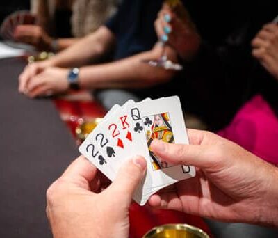 Kädessä on viisi pelikorttia. Käsien takana näkyy tummapintainen pokeripöytä, jossa on punaiset reunat. Takana istuu toinen henkilö.