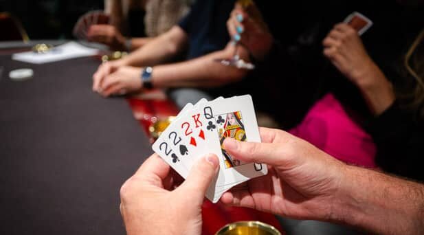 Kädessä on viisi pelikorttia. Käsien takana näkyy tummapintainen pokeripöytä, jossa on punaiset reunat. Takana istuu toinen henkilö.