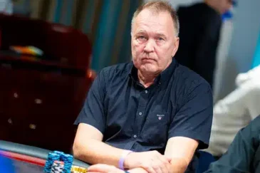 Mies istuu pokeripöydässä. Hänellä on päällään tummansininen t-paita. Hänen edessään pokeripöydällä on erivärisiä pelimerkkejä.