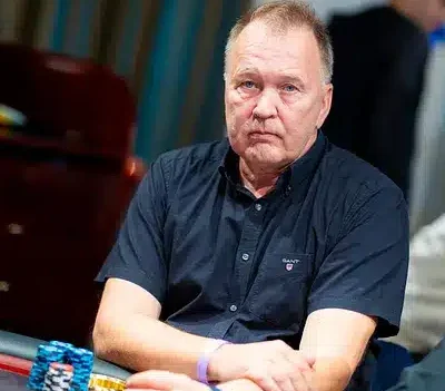 Mies istuu pokeripöydässä. Hänellä on päällään tummansininen t-paita. Hänen edessään pokeripöydällä on erivärisiä pelimerkkejä.