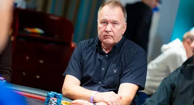 Mies istuu pokeripöydässä. Hänellä on päällään tummansininen t-paita. Hänen edessään pokeripöydällä on erivärisiä pelimerkkejä.