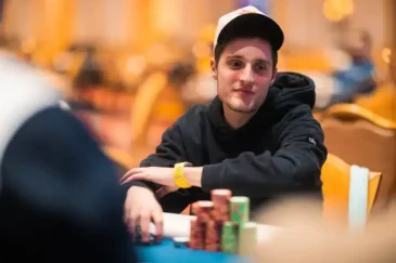 Mies istuu pokeripöydässä. Hänellä on päällään musta huppari ja valkoinen lippis. Edessä pelipöydällä on pelimerkkejä.