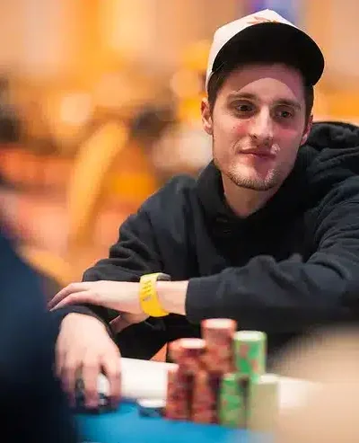 Mies istuu pokeripöydässä. Hänellä on päällään musta huppari ja valkoinen lippis. Edessä pelipöydällä on pelimerkkejä.