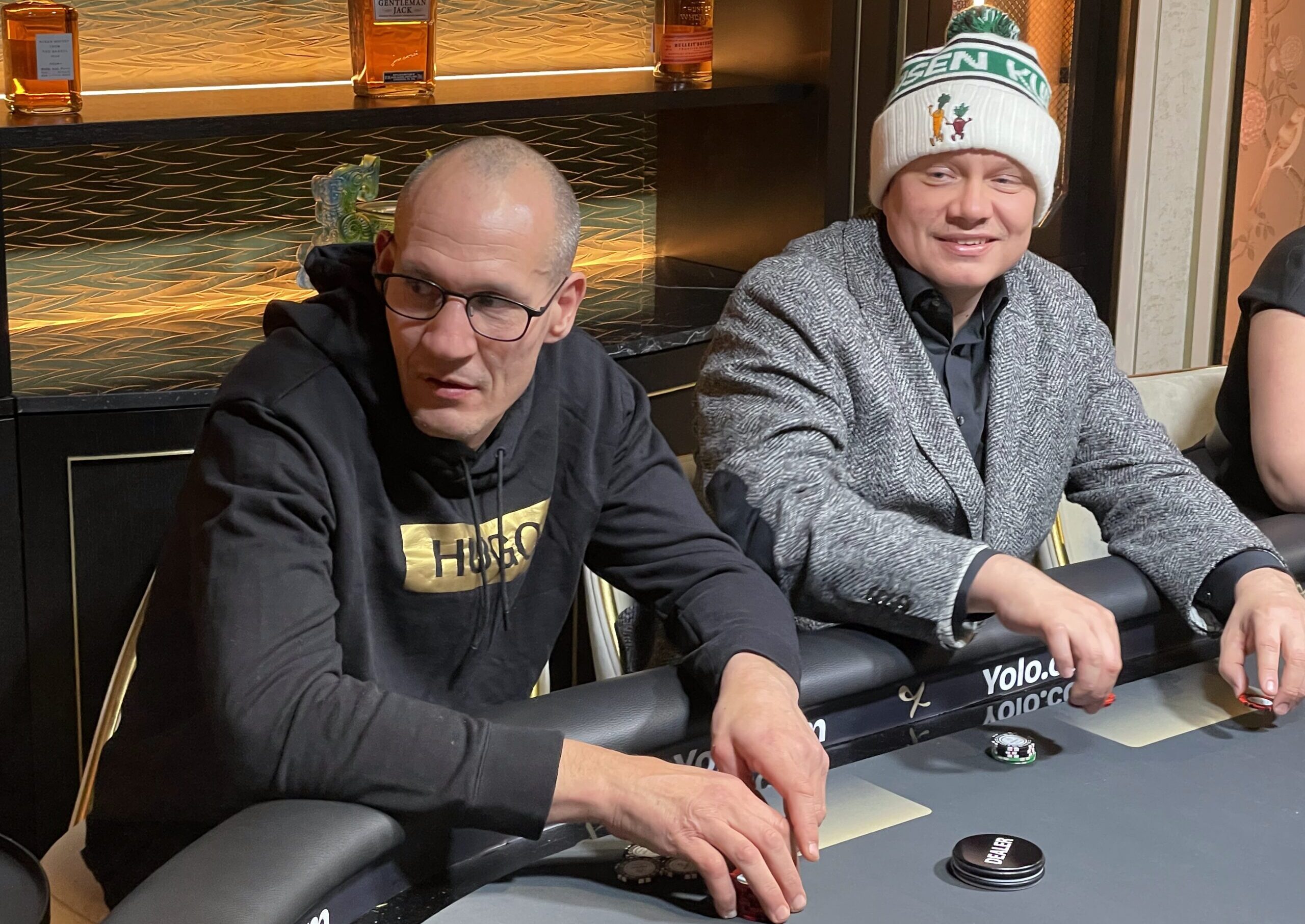 Amin Asikainen ja ilari Sahamies istuvat pokeripöydässä. Pöytä on tumman värinen ja tausta on kultaista väritystä.
