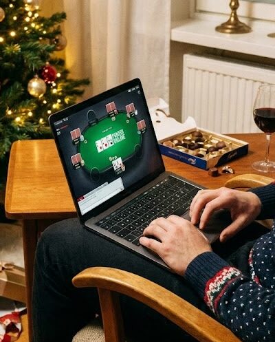 Mies pelaamassa nettipokeria läppäri sylissään joulukuusen äärellä. Pöydällä on punaviiniä ja suklaata