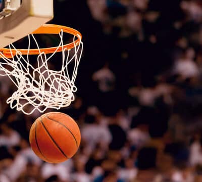 Koripallo on juuri mennyt läpi korirenkaasta ja on laskeutumassa alaspäin. Taustalla on epätarkkoja katsojia.