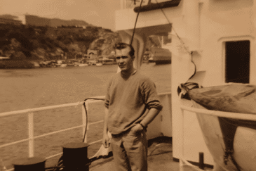 Mies seisoo laivan kannella, taustalla näkyy merta ja kauempana puita. Laivassa on valkoinen aita, miehellä on kädet taskussa.