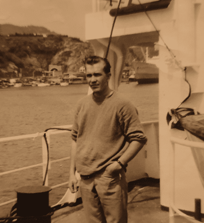 Mies seisoo laivan kannella, taustalla näkyy merta ja kauempana puita. Laivassa on valkoinen aita, miehellä on kädet taskussa.