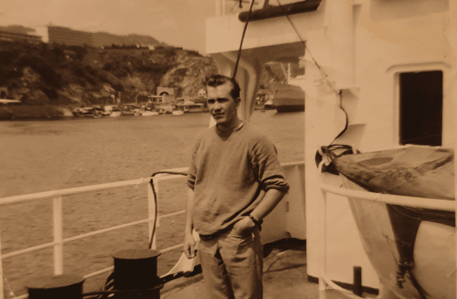 Mies seisoo laivan kannella, taustalla näkyy merta ja kauempana puita. Laivassa on valkoinen aita, miehellä on kädet taskussa.